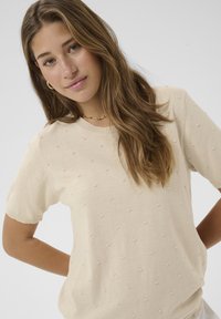 Noorpruunide pikkade juustega noor naine kannab beeži tekstuuriga lühikese varrukaga särki ja õrna kaelakeed, poseerides ühevärvilise taustaga.