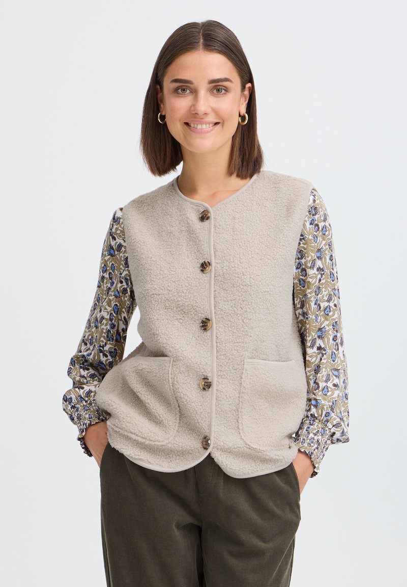 Femme aux cheveux courts et bruns portant des boucles d'oreilles créoles dorées, un gilet polaire beige boutonné, un chemisier à manches longues à motif floral, et un pantalon foncé, souriante, les mains dans les poches.