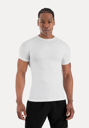 Muskuløs mand med flettet hår iført en tætsiddende hvid kortærmet skjorte og sorte bukser, står mod en ensfarvet lys baggrund.