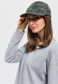 Kapa u kamuflaži s zakrivljenim vizirima, u različitim nijansama zelene i smeđe boje, izrađena od izdržljive tkanine. Nosena s dugim siva košulja.