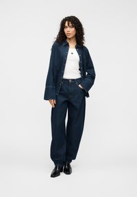 Τζιν πουκάμισο και παντελόνι με φαρδιά πόδια σε σκούρο μπλε χρώμα. Το πουκάμισο διαθέτει κουμπιά, γιακά άνοιγμα· λευκό αμάνικο από μέσα. Μαύρα παπούτσια ολοκληρώνουν την εμφάνιση.