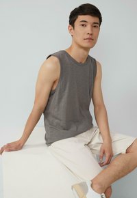 Grijze mouwloze shirt met een ronde halslijn, gecombineerd met lichte shorts. Het model zit op een witte voetstuk en draagt casual sneakers.