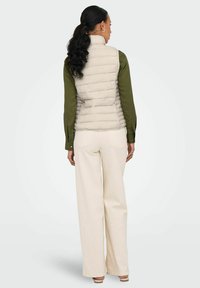 Gilet rembourré beige clair avec des coutures horizontales, fermeture éclair et col montant, porté sur une chemise à manches longues verte et un pantalon crème.