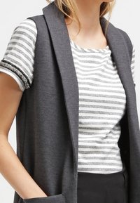 Chaleco gris sin mangas sobre una camiseta de rayas. La camiseta presenta rayas plateadas horizontales y un escote redondo. Tejido suave y casual.