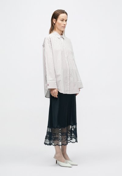 Bielo-červená pruhovaná nadmerná košeľa zapínaná na gombíky v kombinácii s čiernou saténovou sukňou s čipkovaným lemom. Modelka má na sebe biele šľapky na opätku.