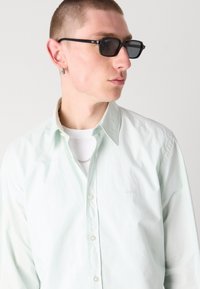 Camisa de botones verde claro hecha de una tela suave, con un pequeño logo blanco, superpuesta sobre una camiseta blanca, con gafas de sol negras puestas.