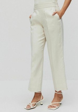 Mujer con pantalones color crema de talle alto y pata ancha, con las manos en los bolsillos y sandalias de tacón blancas de tiras sobre un fondo liso.