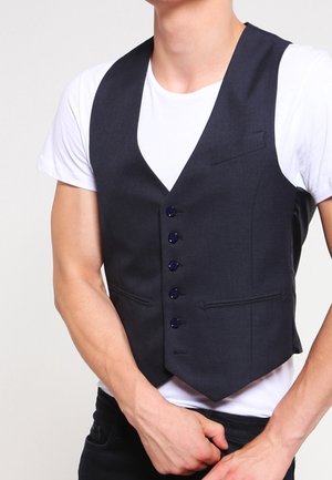 Mężczyzna ubrany w dopasowaną czarną kamizelkę na guziki, założoną na białą koszulkę, z założonymi na wysokości talii skrzyżowanymi ramionami.