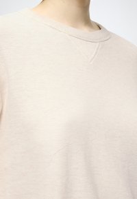 Sweat-shirt beige clair en tissu doux et texturé. Il présente un col rond et des détails de couture subtils avec une légère marque rouge sur le devant.