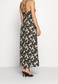 Kwiatowa sukienka maxi z czarnego materiału, z regulowanymi krzyżującymi się ramiączkami, dopasowanym stanikiem i zwiewną spódnicą ozdobioną żółtymi i białymi kwiatami.