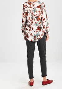 Long-sleeve blouse with a floral pattern in red and green on a light background, paired with tapered dark gray pants and red shoes.