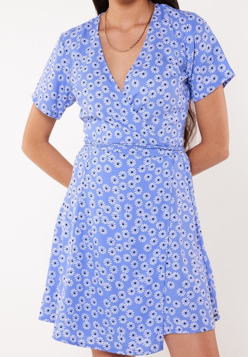 Robe portefeuille bleu à fleurs, avec des manches courtes, un décolleté en V profond et une ceinture à nouer ; décorée de marguerites blanches sur un fond bleu clair.