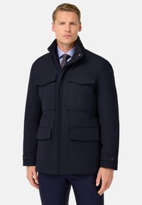 Parka navy con cuello alto, cuatro bolsillos frontales y una textura suave. Diseñada con un puño abotonado para el cierre.