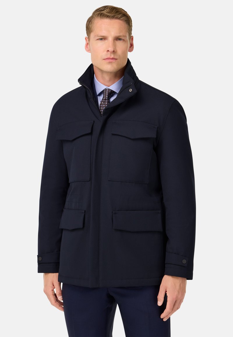Parka navy con cuello alto, cuatro bolsillos frontales y una textura suave. Diseñada con un puño abotonado para el cierre.