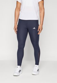 Leggings navy a silhouette aderente realizzati in tessuto elasticizzato, con pannelli in rete sui lati e un logo Adidas bianco sulla coscia sinistra. Abbinati a scarpe da ginnastica bianche.