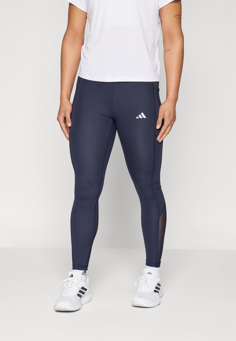 Leggings navy a silhouette aderente realizzati in tessuto elasticizzato, con pannelli in rete sui lati e un logo Adidas bianco sulla coscia sinistra. Abbinati a scarpe da ginnastica bianche.