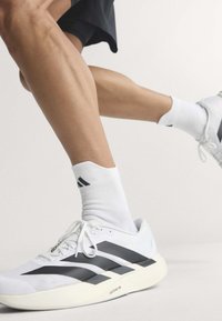Gekreuzte Beine mit weißen Adidas Laufschuhen mit schwarzen Streifen und weißen Socken vor einem einfarbigen hellen Hintergrund.