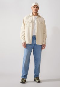 Veste chemise en velours côtelé beige avec deux poches avant, portée sur un t-shirt blanc. Un jean en denim bleu clair et des baskets à motifs complètent le look.