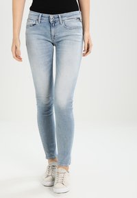 Kobieta w jasnoniebieskich obcisłych dżinsach i beżowych trampkach, stojąca na jednolitym jasnoszarym tle.