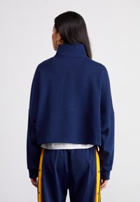 Sudadera recortada de color azul marino con un cuello alto, que presenta una tela texturizada y un corte relajado. Combinada con pantalones azules con franjas laterales amarillas.