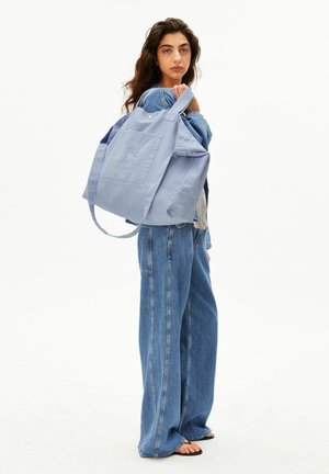 Woman with wavy dark hair wearing denim jacket and wide-leg jeans carries large light blue tote bag over shoulder, standing against plain background.