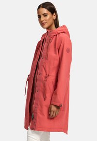 Coral parka with hood, drawstring waist, front zipper, and visible hardware. Features a logo patch on the left sleeve. Soft, smooth fabric.