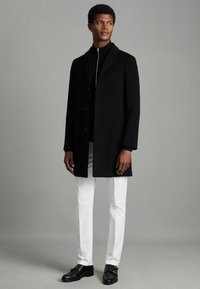 Cappotto di lana nero con collo a scoprire, tre bottoni, abbinato a un maglione nero a collo alto, pantaloni bianchi e scarpe in pelle nere.