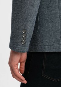 Grå vävd jackärm med fyra mörka knappar, med subtil textur och skräddarsydd passform. Hand placerad mot mörkt tyg.