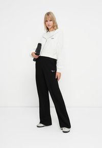 Gekürzte weiße Zip-Up-Hoodie mit gerippter Textur, kombiniert mit lockeren schwarzen Hosen mit Logo. Das Model hält eine kleine schwarze Tasche.