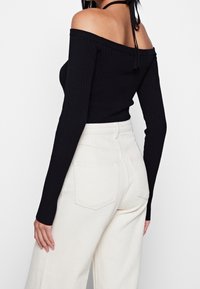 Schwarzes geripptes Off-Shoulder-Oberteil mit langen Ärmeln, kombiniert mit hochgeschnittenen, hellbeigen, weit geschnittenen Hosen mit Gesäßtaschen.