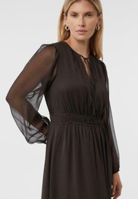 Braunes, langärmliges Kleid mit tailliertem Schnitt, aus fließendem Stoff. V-Ausschnitt mit Bindeband und transparenten Ärmeln.