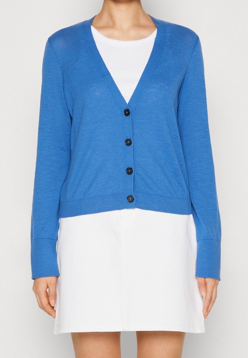 Femme portant un cardigan bleu boutonné par-dessus un haut blanc avec une jupe blanche, visible des épaules jusqu'à mi-cuisse sur un fond uni.