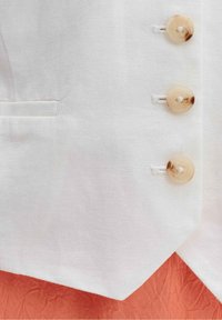 Gros plan d’un gilet en tissu blanc avec trois boutons marron et une petite poche fendue sur un fond en tissu orange texturé.