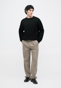Zwarte geribbelde gebreide trui, beige gestructureerde broek en zwarte schoenen. Model staat rechtop tegen een effen achtergrond. Krullend haar zichtbaar.