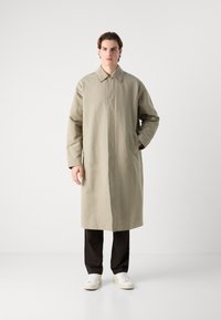 Beige lange jas met een kraag, zijzakken en een rechte snit, gecombineerd met donkere broeken en witte sneakers op een effen achtergrond.