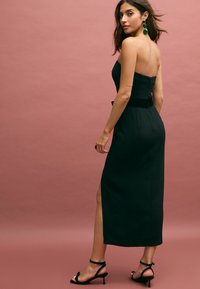 Svart off-shoulder klänning med figurnära bodice, smal midja och sidoslits. Bärs med svarta klacksandaler, mot en rosa bakgrund.