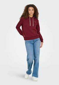 Vinrød hoodie med snoretræk, udstyret med en lille hjertebrodering og ribbet kant. Parret med lyseblå flagrende jeans og hvide sneakers.
