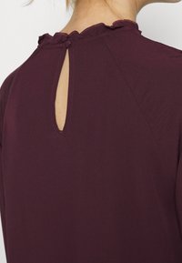 Blusa bordeaux con scollo arricciato, apertura a goccia sul retro e texture di tessuto liscia e leggera.