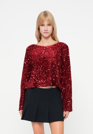 Top de manga longa vermelho com lantejoulas, com decote arredondado e corte cropped, combinado com uma saia preta plissada. Textura suave e acabamento brilhante.