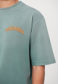 Camiseta de algodón verde claro con bordado naranja "DSQUARED2" en la parte superior izquierda, mangas cortas y escote redondo.