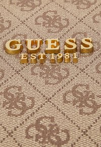 Letras doradas "GUESS" y "EST 1981" en relieve sobre un fondo texturizado de color marrón claro con patrón. Los patrones presentan diseños entrelazados.