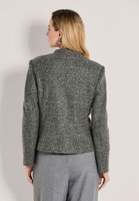 Blonde vrouw met de rug naar de camera, gekleed in een gestructureerde grijze wollen blazer met gewatteerde schouders en lichtgrijze op maat gemaakte broek.