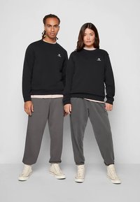 Sudaderas negras combinadas con pantalones de chándal grises. Ambos tienen puños elásticos y presentan diseños minimalistas con logotipos. El calzado casual completa el conjunto.