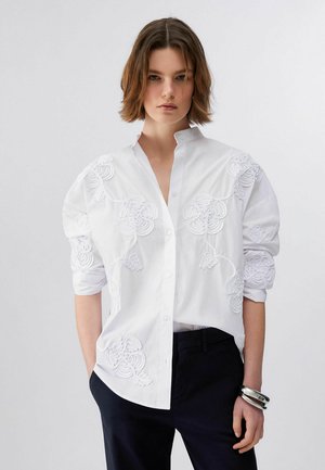 Giovane donna con capelli castani corti che indossa una camicia bianca ricamata con bottoni e pantaloni neri, posando con una mano in tasca.