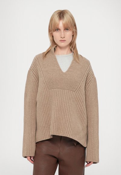 Beige gestrickter Pullover mit V-Ausschnitt und gerippter Struktur. Hat überschnittene Schultern und eine lockere Passform. Trägt ihn über einem hellgrauen Oberteil und braunen Hosen.
