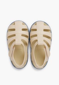 Light beige rubber sandals with structured straps, open sides, and a textured blue sole. Featuring a label on the heel.