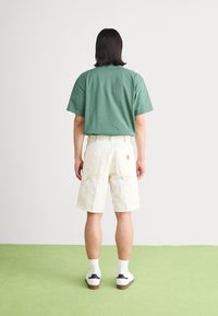 T-shirt en coton vert, shorts cargo blancs avec de multiples poches, associés à des baskets blanches et des chaussettes mi-mollet, vus de dos.