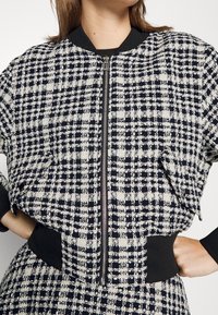 Femme portant une veste zippée noir et blanc à motif pied-de-poule, avec des poignets et une taille noirs, les mains posées sur les hanches.