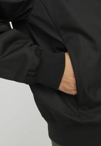 Chaqueta bomber negra con una textura suave y brillante. Presenta puños acanalados y una mano en un bolsillo lateral, destacando su diseño casual.