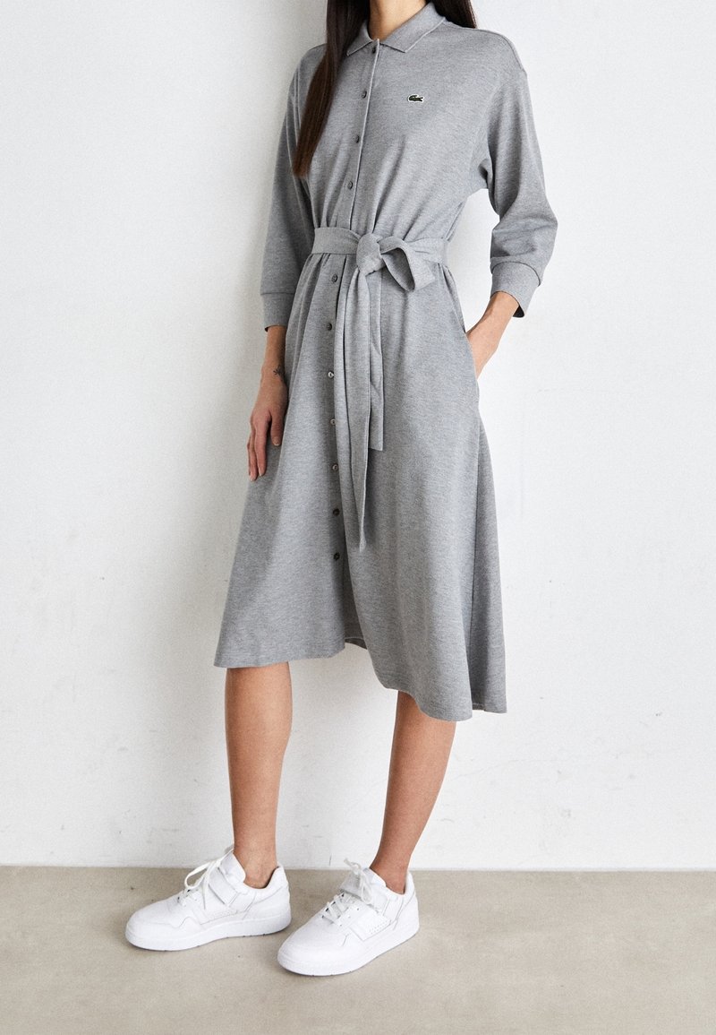 Femme portant une robe grise à manches longues, boutonnée, avec une ceinture à la taille et des baskets blanches, debout contre un mur blanc uni.